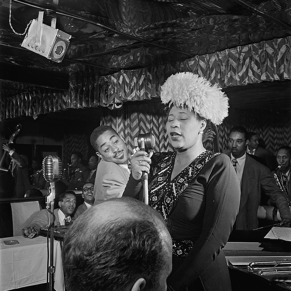 Black and white photograph of Ella Fitzgerald performing in a New York City club, wearing a dark dress and small feathered cap. Musicians Dizzy Gillespie, Ray Brown, Milt Jackson, and Timmie Rosenkrantz appear in the background, 1947.]