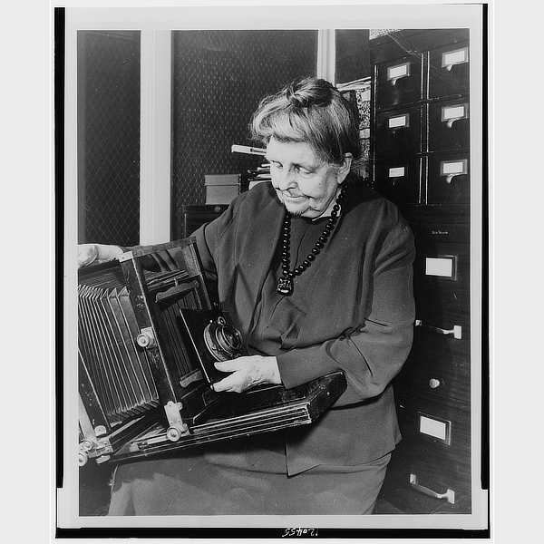 Black and white image of photographer Frances Benjamin Johnston, seated and looking down at a large format camera she is holding.]