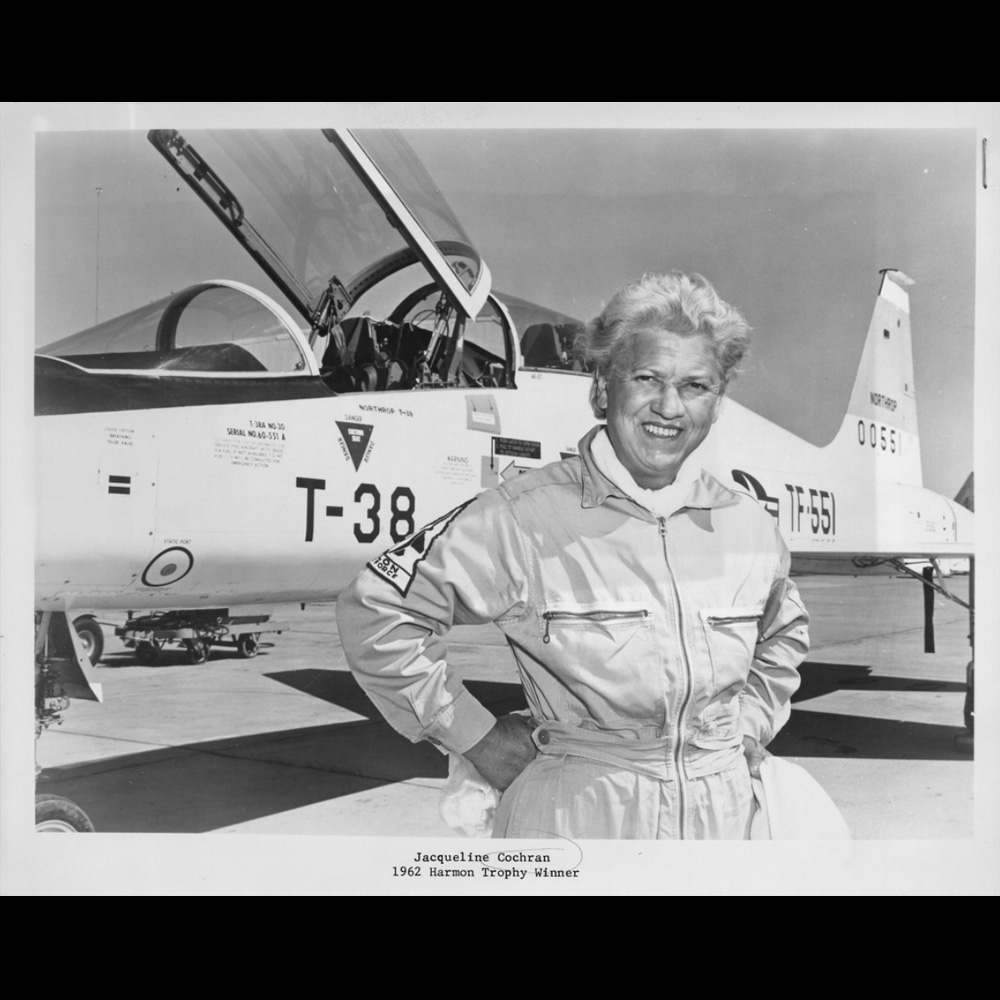 Black and white photograph of pilot Jacqueline Cochran in a flight suit with arms akimbo, standing in front of a U.S. Air Force jet.]