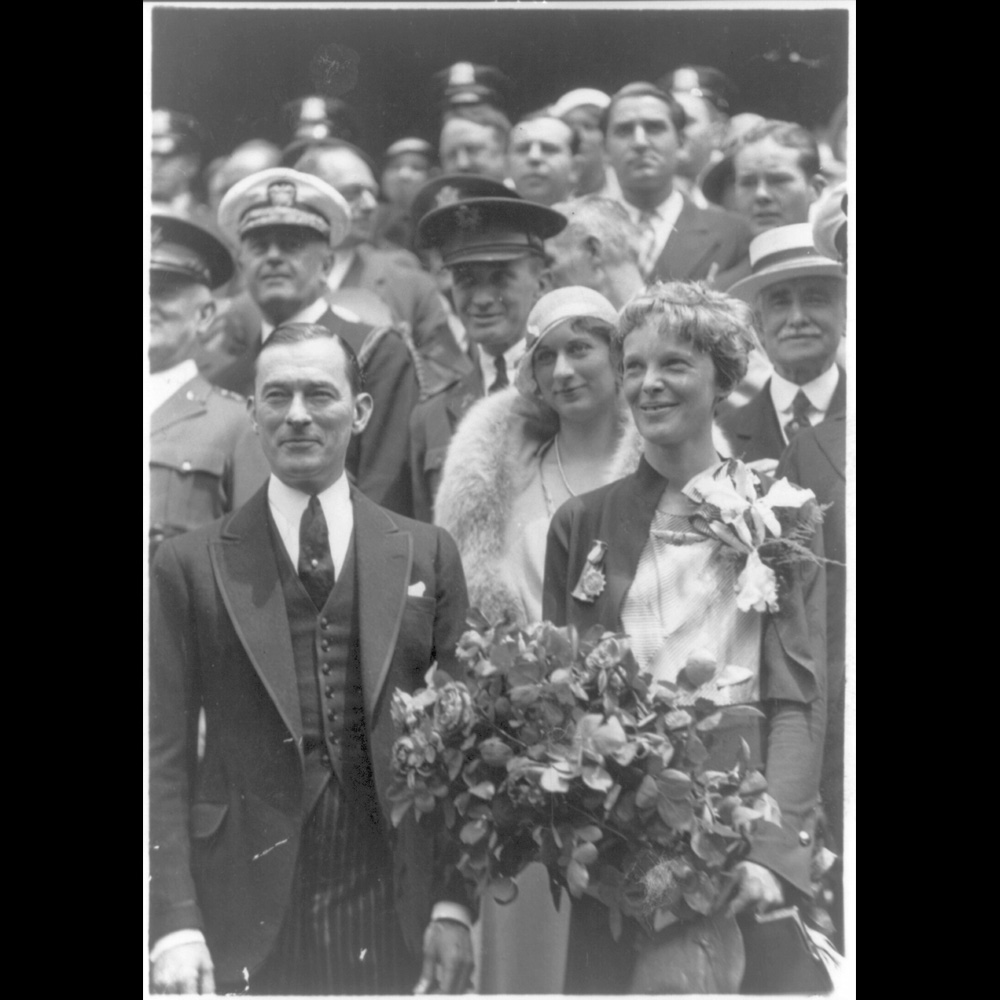 Black and white photograph of Amelia Earhart, holding a large bouquet of flowers, standing with Mayor Jimmy Walker of New York City.]