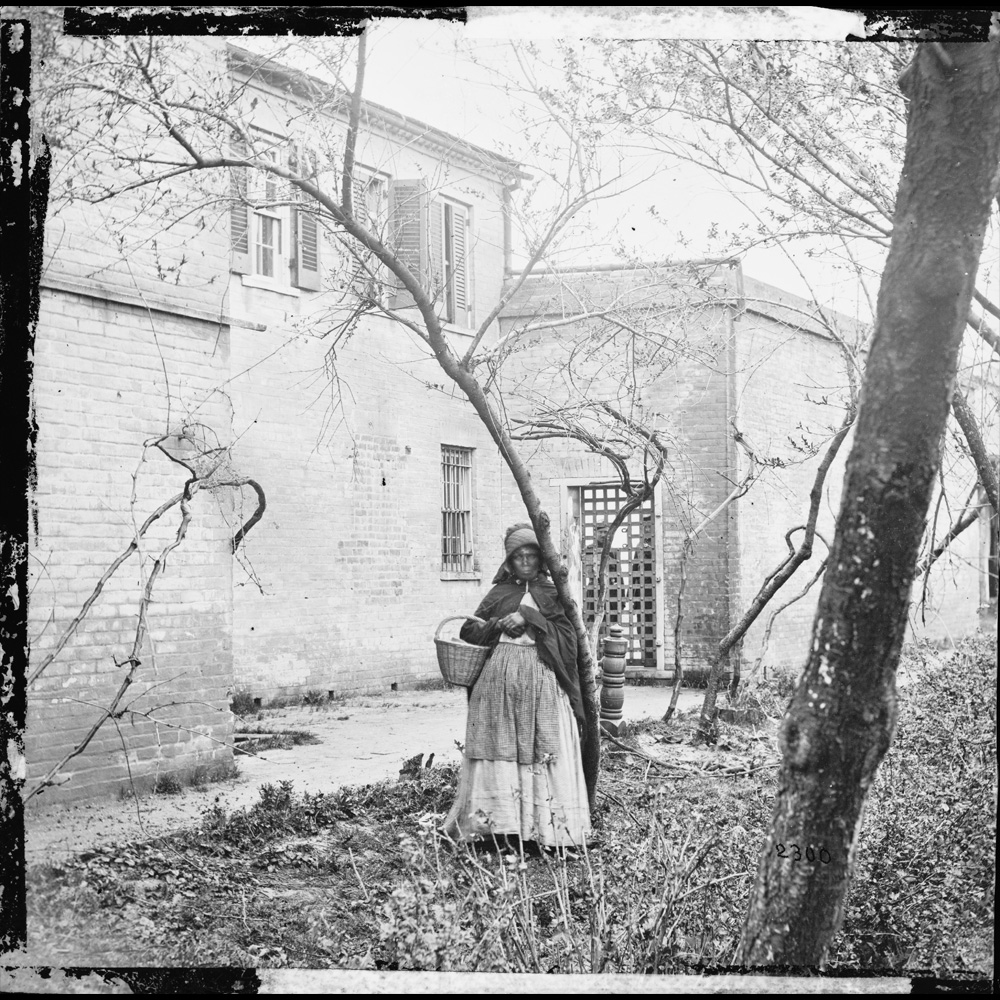 African American woman dressed in a cloak, bonnet and apron, holding a wicker basket standing outside the building known as the Slave Pen in Alexandria, Virginia. Taken sometime between 1861 and 1869.]