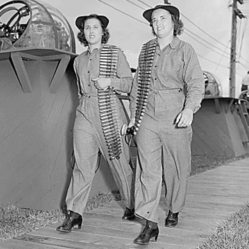 Black and white photograph of two WAVES machine gunnery instructors wearing fatigue jumpsuits and carrying lengths of machine gun ammunition over their shoulders.]