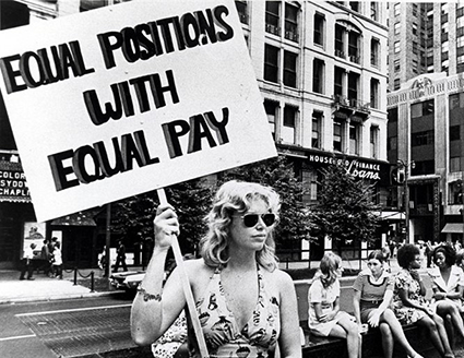Black and white photograph of a young woman in a printed summer dress and sunglasses holding a protest sign which reads &ldquo;Equal Positions With Equal Pay&rdquo;