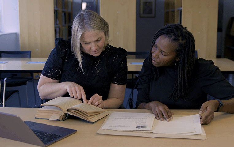 Two women reviewing historical documents in a Library setting.