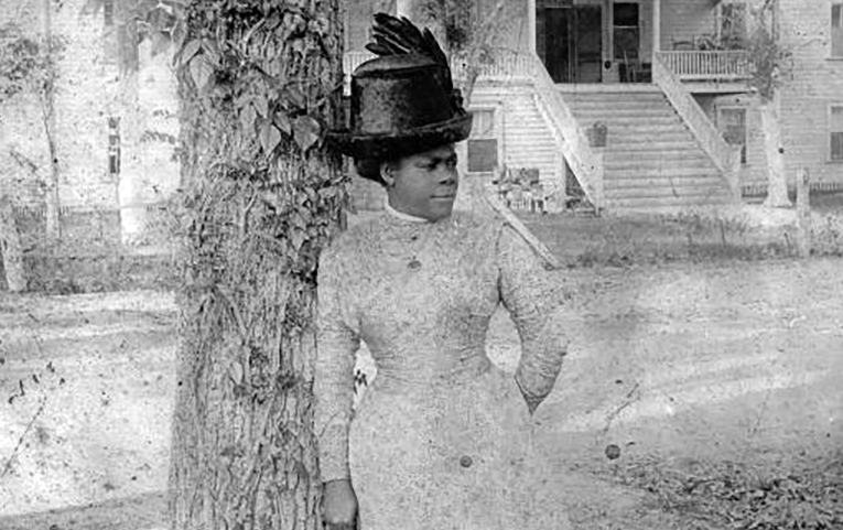 Black and white image of Mary McLeod Bethune standing beneath a tree wearing a pale, lace dress and a tall black hat with feathers on the left side.