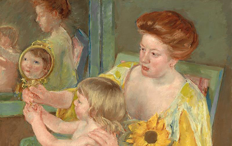 A red haired woman wearing a long, green and yellow dress with a large sunflower pinned to the breast sits in a green wooden chair with a small child seated on her lap. The child looks at her reflection in the woman’s hand mirror, while both subjects are reflected in a nearby wall mirror.
